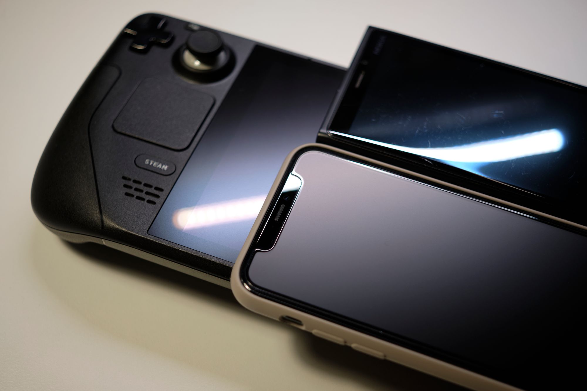 Showing a Steam Deck, with an iPhone with a matte screen protector (11 Pro) and a old Nokia phone (Lumia 920) on top. And all 3 devices are angled to try to reflect a bar of LED light, in which it shows different level of reflectiveness. 