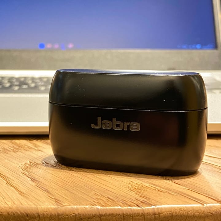 Jabra Elite 75t case on a wooden desk, in front of a silver laptop